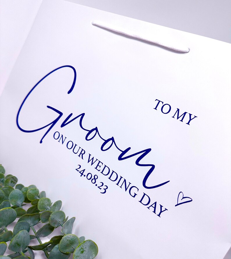 Puede incluir: Una bolsa de regalo blanca con una inscripci&oacute;n azul que dice "To my Groom on our Wedding Day 24.08.23" con un peque&ntilde;o s&iacute;mbolo de coraz&oacute;n.