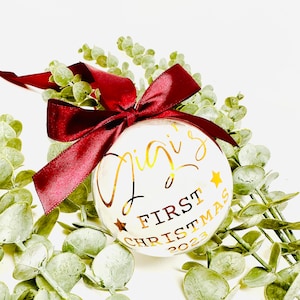 Puede incluir: Adorno navide&ntilde;o blanco con letras doradas que dicen "Gigi's First Christmas 2023". El adorno est&aacute; decorado con estrellas doradas y est&aacute; atado con una cinta color burdeos.