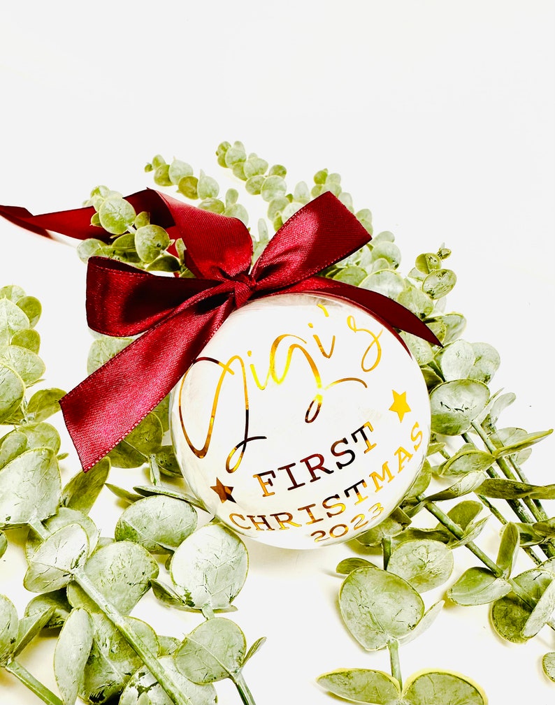 Puede incluir: Adorno navide&ntilde;o blanco con letras doradas que dicen "Gigi's First Christmas 2023". El adorno est&aacute; decorado con estrellas doradas y est&aacute; atado con una cinta color burdeos.