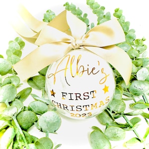 Puede incluir: Adorno navide&ntilde;o blanco con letras doradas que dicen "Albie's First Christmas 2023". El adorno est&aacute; decorado con estrellas doradas y atado con una cinta dorada.