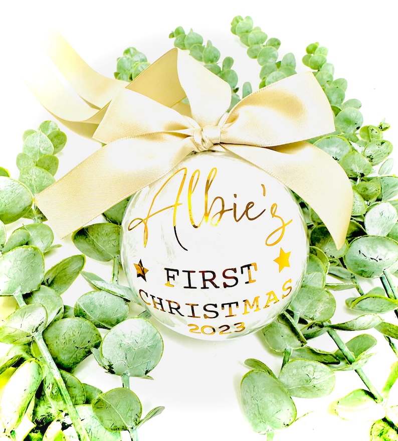 Puede incluir: Adorno navide&ntilde;o blanco con letras doradas que dicen "Albie's First Christmas 2023". El adorno est&aacute; decorado con estrellas doradas y atado con una cinta dorada.