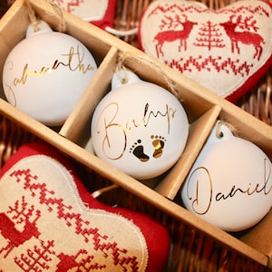 Puede incluir: Adornos navideños blancos con letras doradas, presentados en una caja de madera. Uno dice "Samantha", otro "Baby" con huellas y el tercero "Daniel". También se ven adornos en forma de corazón con diseños rojos y blancos.