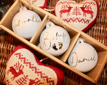 Puede incluir: Adornos navideños blancos con letras doradas, presentados en una caja de madera. Uno dice "Samantha", otro "Baby" con huellas y el tercero "Daniel". También se ven adornos en forma de corazón con diseños rojos y blancos.