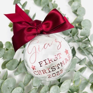 Puede incluir: Un adorno de Navidad de vidrio blanco con un lazo de cinta color burdeos. El adorno est&aacute; decorado con plumas blancas y tiene el texto "Gia's First Christmas 2022" en letras doradas.