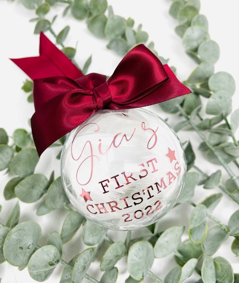 Puede incluir: Un adorno de Navidad de vidrio blanco con un lazo de cinta color burdeos. El adorno est&aacute; decorado con plumas blancas y tiene el texto "Gia's First Christmas 2022" en letras doradas.