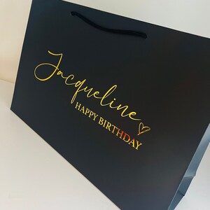 May include: Black gift bag with a gold inscription that reads "Jacqueline Happy Birthday" with a gold heart.