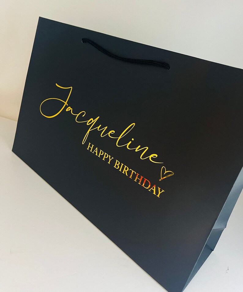 May include: Black gift bag with a gold inscription that reads "Jacqueline Happy Birthday" with a gold heart.