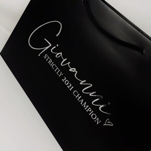 May include: Black shopping bag with a white logo that says "Giovanni" and "Strictly 2021 Champion" with a heart.