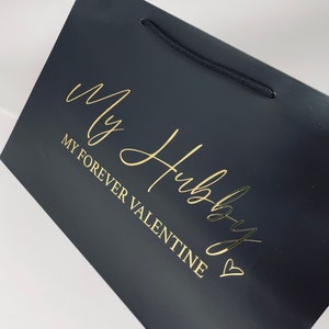 May include: Black gift bag with a gold inscription that reads "My Hubby My Forever Valentine" with a heart symbol.