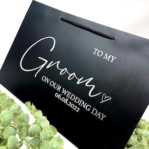 Puede incluir: Bolsa de regalo negra con una inscripci&oacute;n blanca que dice "To My Groom On Our Wedding Day 06.08.2022" con un peque&ntilde;o s&iacute;mbolo de coraz&oacute;n.