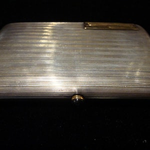 Könnte beinhalten: Ein silbernes Zigarettenetui mit einem goldfarbenen Band und einem strukturierten, gestreiften Design.
