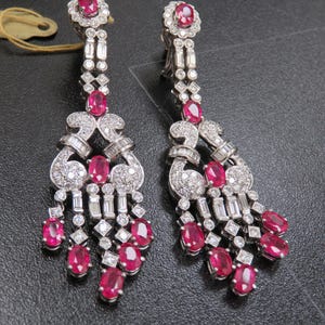 Puede incluir: Un par de pendientes colgantes con rubíes y diamantes. Los pendientes tienen un diseño floral en la parte superior y una disposición en cascada de gemas. Los rubíes son de corte ovalado.