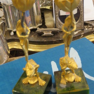 Pode incluir: Dois castiçais decorativos dourados em forma de tulipa estilizada com um sapo na base. Cada castiçal está sobre uma base quadrada verde.