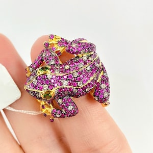 May include: A gold ring featuring a detailed frog design, set with pink and white gemstones. The frog's eyes are green.