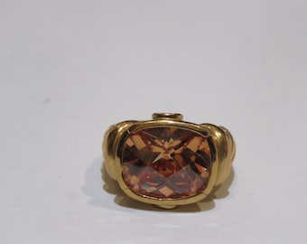 Puede incluir: Un anillo dorado con una gran gema facetada de color marrón anaranjado. El anillo tiene una banda redondeada y texturizada y un engaste cuadrado. La gema tiene múltiples facetas.