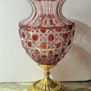 May include: A large, ornate, ruby red glass vase with a cut glass pattern. The vase has a gold metal base with a decorative design.
