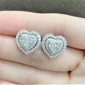 Może przedstawiać: Para kolczyków w kształcie serca z diamentem w podwójnej oprawie halo. Kolczyki są wykonane z białego złota i mają błyszczący kamień centralny.