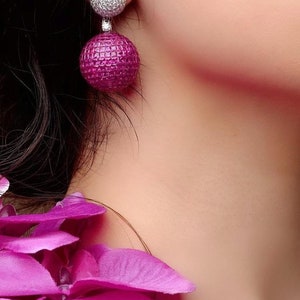Pode incluir: Um par de brincos pendentes com um design rosa e branco. A parte superior do brinco é uma bola branca coberta de pequenas pedras transparentes. A parte inferior do brinco é uma bola rosa maior com uma superfície texturizada.