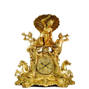 May include: A gold-toned clock with a Roman numeral face and an intricate design featuring a woman holding a seashell, two cherubs blowing trumpets, and two seahorses.
