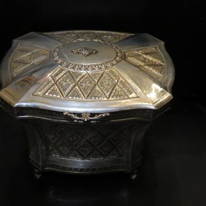 Könnte beinhalten: Silberfarbene, achteckige Schmuckschatulle mit aufwendigen Details. Der Deckel hat ein zentrales ovales Design und rautenförmige Muster. Die Schatulle hat einen dekorativen Verschluss und steht auf kleinen Füßen, was eine Vintage-Ästhetik zeigt.