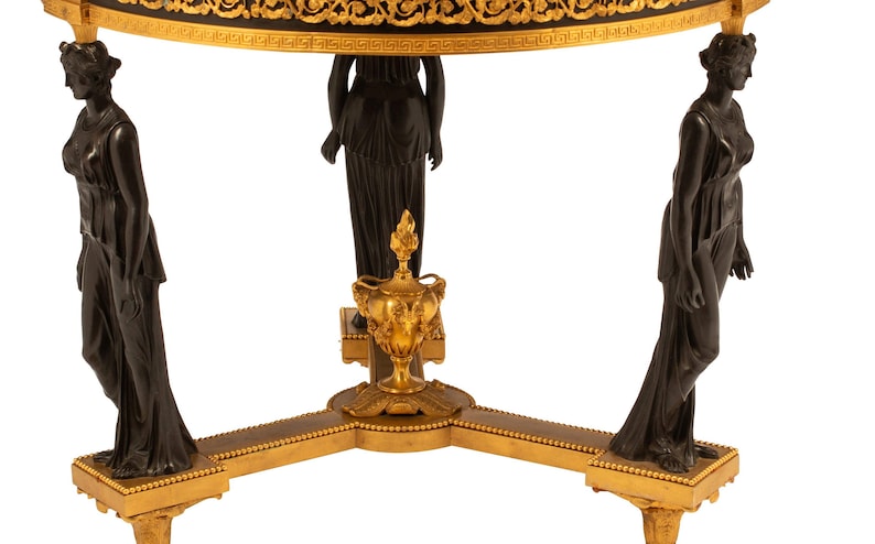 May include: A black and gold table with three legs, each featuring a bronze statue of a woman. The table top has a black and gold design.