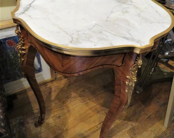May include: A small, oval-shaped table with a white marble top and a gold trim. The table has ornate, carved legs and a wooden base with a marquetry design.
