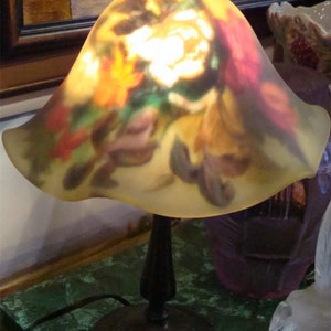 Könnte beinhalten: Eine Tischlampe mit einem floral gemusterten Glasschirm. Der Schirm ist hellgelb mit Rot-, Grün- und Lilatönen. Der Lampenfuß ist dunkelbraun.