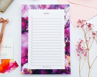 May include: A pink and purple abstract art notepad with a white lined section for writing notes. The notepad has a gold border and the text "NOTES" and "EMMA GODFREY ART" printed on it.