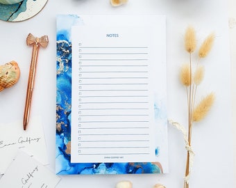 May include: A white notepad with a blue and gold abstract design. The notepad has a checklist with the word "NOTES" at the top. The notepad is on a white surface with other decorative items.