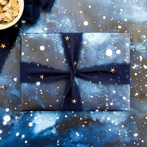 May include: A wrapped gift with a celestial theme, featuring navy blue wrapping paper with white speckles and a watercolor effect. The gift is tied with a navy blue ribbon and adorned with gold star embellishments. A small bowl of gold confetti sits nearby.