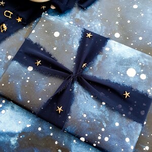 May include: A wrapped gift with a blue and white galaxy-themed paper, adorned with a dark blue ribbon and gold star embellishments. A small white bowl holds gold confetti. The overall aesthetic is celestial and festive.