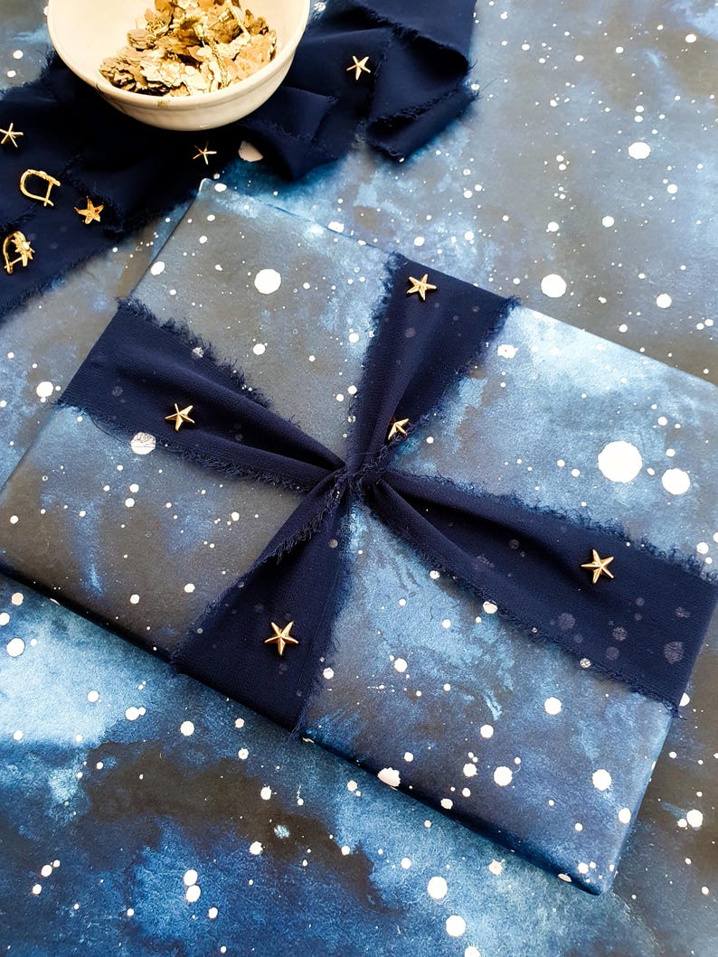 May include: A wrapped gift with a blue and white galaxy-themed paper, adorned with a dark blue ribbon and gold star embellishments. A small white bowl holds gold confetti. The overall aesthetic is celestial and festive.