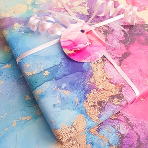 May include: Two sheets of wrapping paper with a blue, pink and purple watercolour design with gold flecks. The paper is tied with a white ribbon and a pink circle tag.