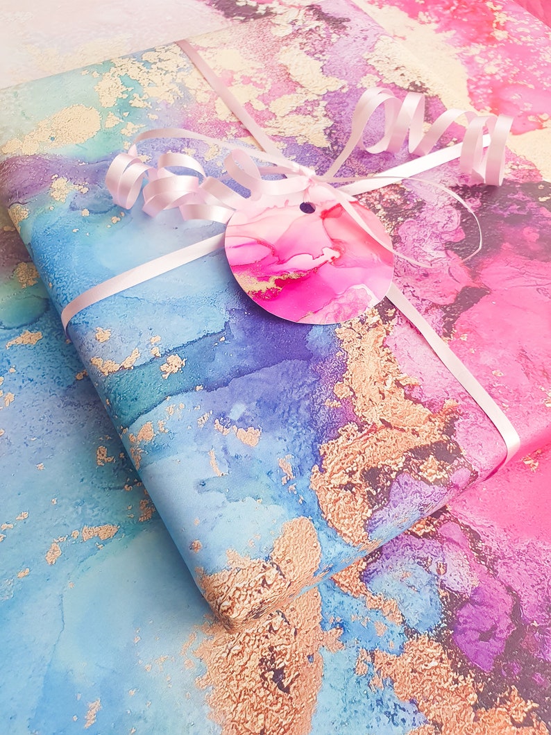 May include: Two sheets of wrapping paper with a blue, pink and purple watercolour design with gold flecks. The paper is tied with a white ribbon and a pink circle tag.