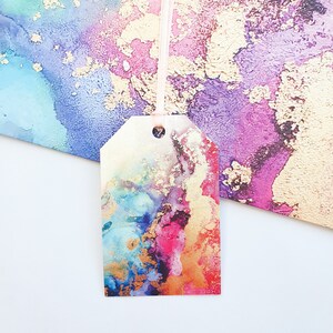 May include: A colourful gift tag with a watercolour design featuring shades of blue, pink, orange, and gold. The tag has a hole punched in the top for a ribbon or string.