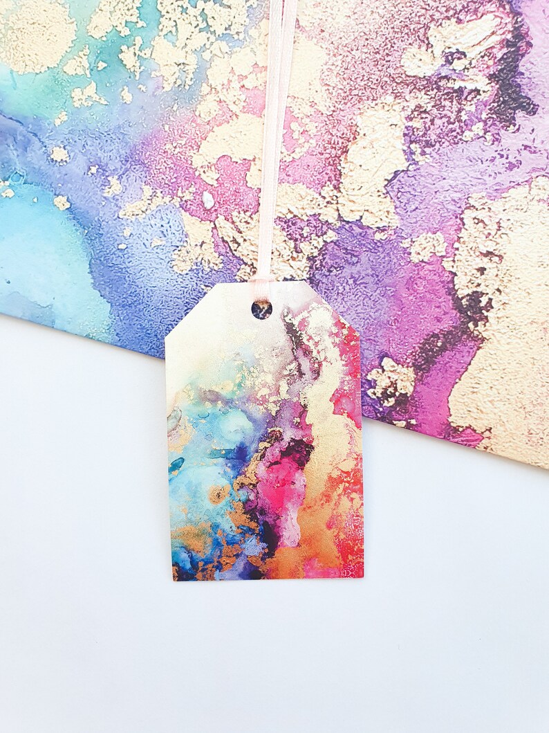 May include: A colourful gift tag with a watercolour design featuring shades of blue, pink, orange, and gold. The tag has a hole punched in the top for a ribbon or string.