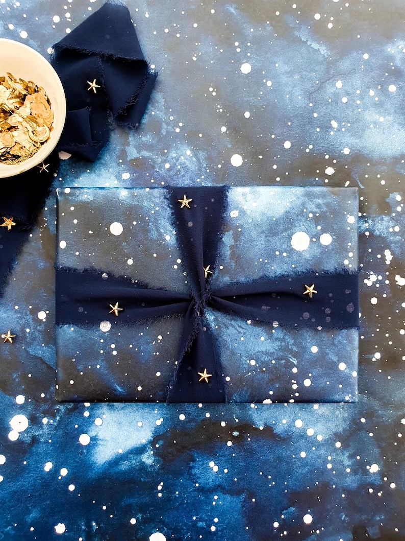 May include: A wrapped gift with a celestial theme, featuring navy blue wrapping paper with white speckles and a watercolor effect. The gift is tied with a navy blue ribbon and adorned with gold star embellishments. A small bowl of gold confetti sits nearby.