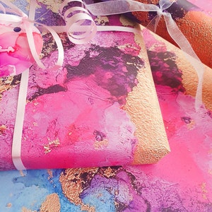 May include: Pink, blue and gold abstract wrapping paper with a pink ribbon and a white bow. The paper has a textured, watercolour-like design.