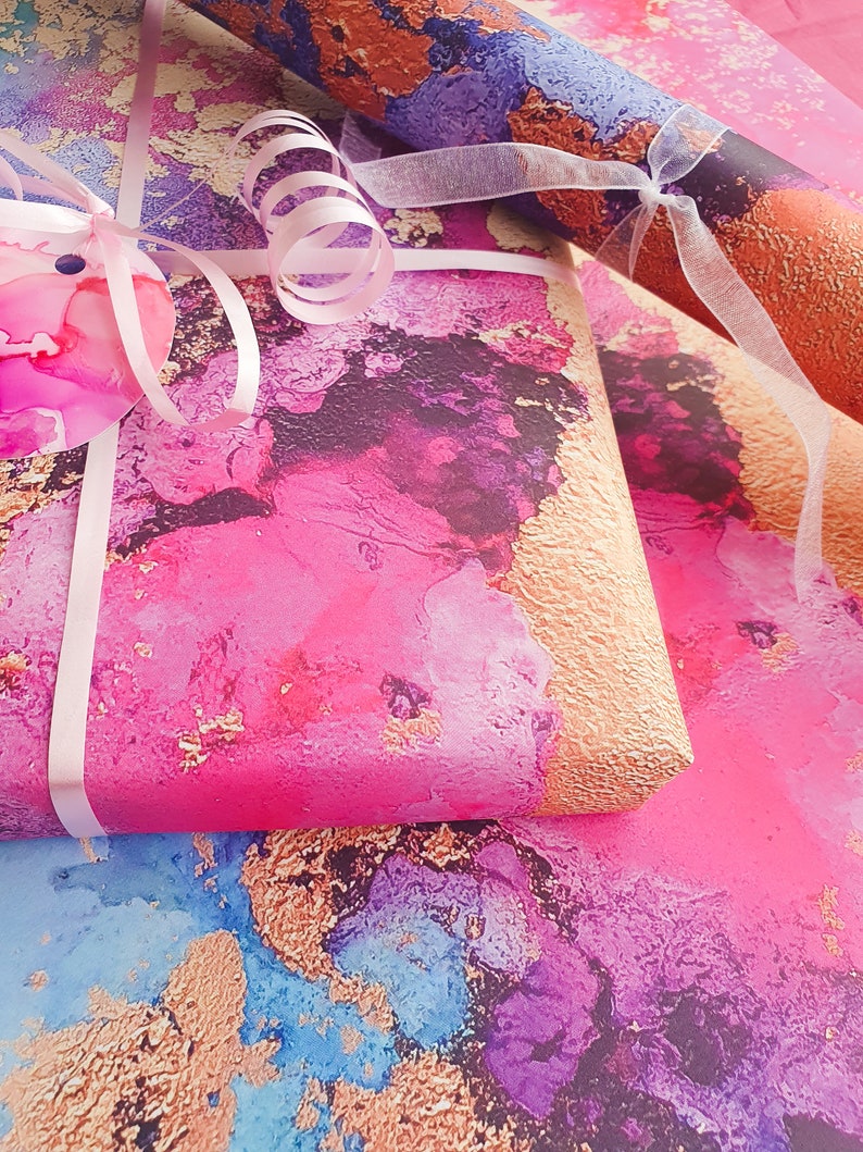 May include: Pink, blue and gold abstract wrapping paper with a pink ribbon and a white bow. The paper has a textured, watercolour-like design.