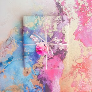 May include: A gift wrapped in colourful paper with gold accents. The paper features blue, pink and purple hues with a marbled effect. The gift is tied with a pink ribbon and a circular tag.