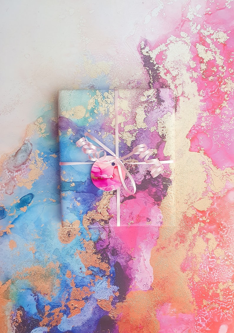 May include: A gift wrapped in colourful paper with gold accents. The paper features blue, pink and purple hues with a marbled effect. The gift is tied with a pink ribbon and a circular tag.
