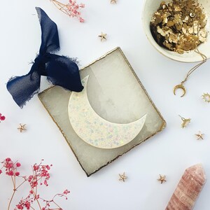 May include: A glittery moon-shaped ornament with a gold-edged square base and a navy blue ribbon. The image also features gold star charms, a crescent moon necklace, and a pink crystal on a white background.