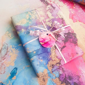 May include: A gift wrapped in blue, pink and purple marbled paper with gold accents. The paper has a watercolour effect and is tied with a white ribbon and a pink bow. A pink circle tag with a pink and white marbled design is attached to the ribbon.