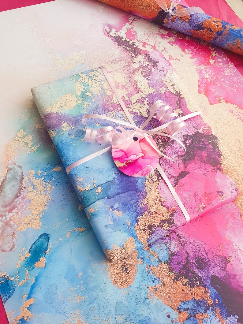 May include: A gift wrapped in blue, pink and purple marbled paper with gold accents. The paper has a watercolour effect and is tied with a white ribbon and a pink bow. A pink circle tag with a pink and white marbled design is attached to the ribbon.