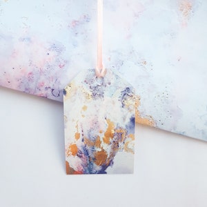 May include: A white gift tag with a watercolour design featuring shades of purple, pink, and gold. The tag is tied with a pink ribbon.