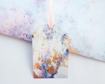 May include: A white gift tag with a watercolor design featuring shades of purple, pink, and gold. The tag is tied with a pink ribbon.