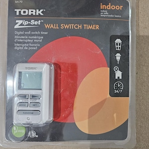 Puede incluir: Un temporizador de interruptor de pared digital TORK Zip-Set en un paquete de plástico transparente. El temporizador tiene una cara blanca con una pantalla digital y varios botones. El embalaje incluye las palabras "indoor" y "24/7".