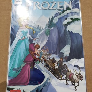 May include: A comic book cover featuring the Disney Frozen characters Elsa and Anna. Elsa is wearing a light blue gown and Anna is wearing a pink and blue dress. The cover features the title "Disney Frozen" in large, bold letters. The background is a snowy mountain scene.