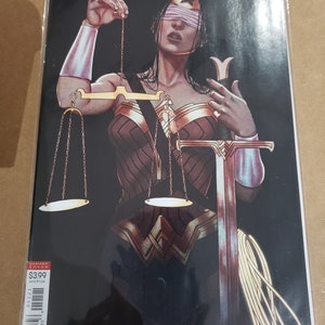 May include: Wonder Woman comic book cover featuring a woman wearing a blindfold and holding scales of justice and a sword. The cover is black with gold accents and the text "Wonder Woman 51 Orlando Braga Fajardo Jr" is at the bottom.