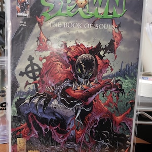 以下が含まれることがあります： 「Spawn: The Book of Souls」のコミック表紙。赤いマントとトゲのある鎧を着たSpawnが墓地を背景に描かれています。タイトルは緑色で、スカルのグラフィックが特徴です。コミックは保護スリーブに入っています。